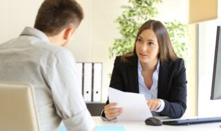 Young woman in a suit speaking to a man at a desk while holding a piece of paper