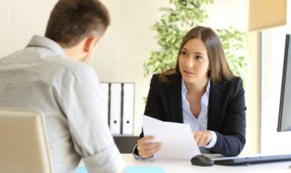 Young woman in a suit speaking to a man at a desk while holding a piece of paper