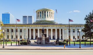 Front view of a neoclassical government building with a circular dome, tall columns, flags, and a statue in the front plaza.