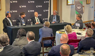Panel of five men in suits seated at a long table on a stage with an NFIB backdrop, speaking into microphones.