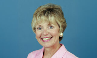 Smiling woman with short blonde hair in a pink blazer and pearl earrings against a blue background.