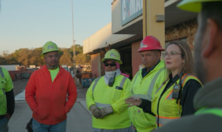 Group of construction workers in high-visibility vests and hard hats standing in a circle outside a building, listening to a speaker.