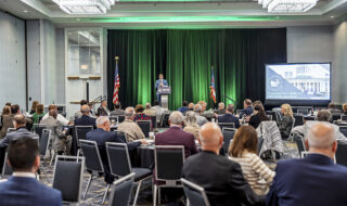 Speaker at a podium on a stage with green curtains and NFIB logo, addressing a conference audience in a banquet hall. American flag visible on the left.