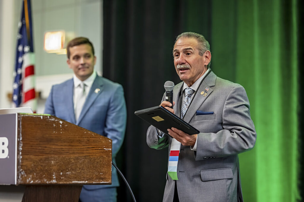 NFIB Small Business Day at the Capital - NFIB Man in a gray suit speaks into a handheld microphone at a wooden podium, holding a tablet; another man in a blue suit stands in the background with an American flag nearby.
