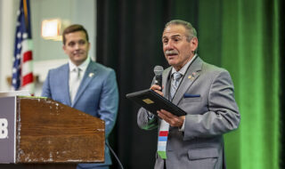 Man in a gray suit speaks into a handheld microphone at a wooden podium, holding a tablet; another man in a blue suit stands in the background with an American flag nearby.