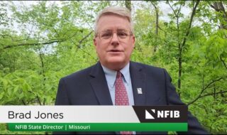 Brad Jones, NFIB Missouri State Director, wearing a dark suit and red tie, speaking outdoors with green trees behind; NFIB banner visible at the bottom.