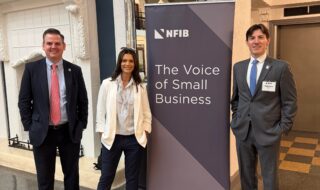 Three professionals posing beside an NFIB banner reading 'The Voice of Small Business' in an ornate indoor lobby.