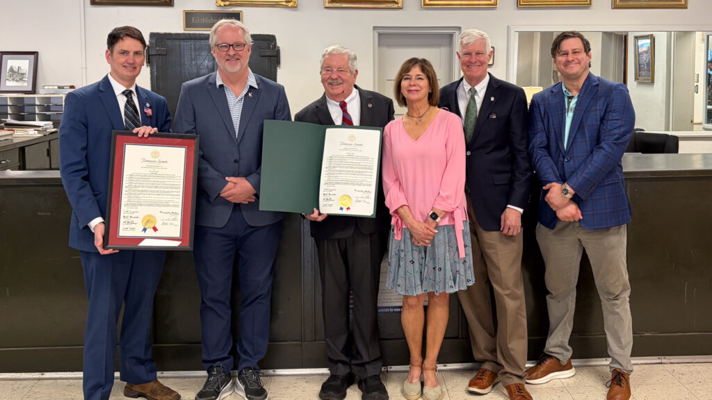 IMG_5251 - NFIB Six adults in formal attire pose together; two men hold large framed certificates, while a woman in a pink blouse stands beside them in a reception-like room.