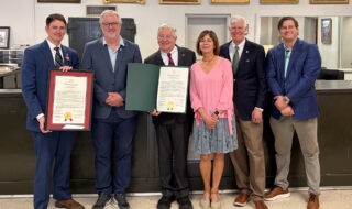 Six adults in formal attire pose together; two men hold large framed certificates, while a woman in a pink blouse stands beside them in a reception-like room.