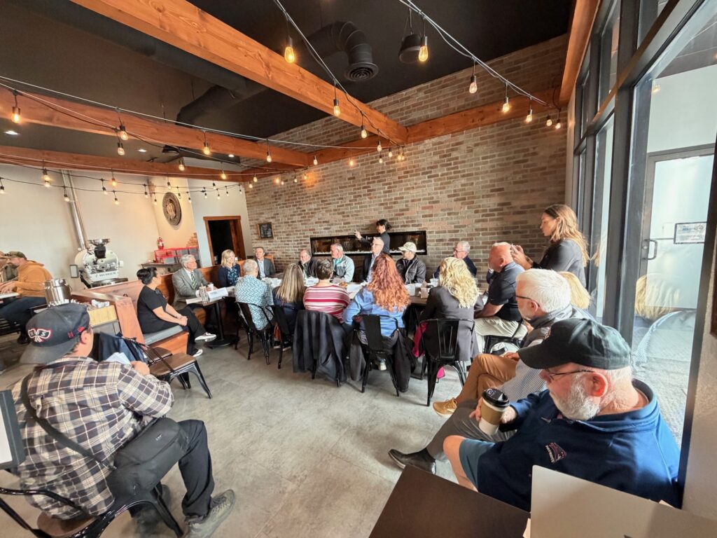 A large group of people seated around long tables in a cafe-style meeting space with brick walls and hanging string lights.