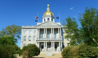 Front view of a grand government building with a gold dome, tall columns, and a statue in front beneath a clear blue sky.