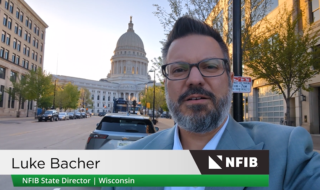 Portrait of Luke Bacher wearing a suit and glasses, recording on a city street with a domed capitol building in the background; NFIB banner shows his name and title (Wisconsin).
