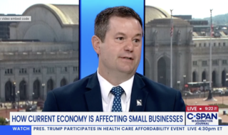 Man in a dark suit and blue polka-dot tie speaks during a live C-SPAN Washington Journal interview, with a cityscape backdrop visible behind him.