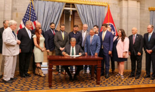 Man signs a document at a wooden desk while a group of officials observes in a formal room with flags behind him.
