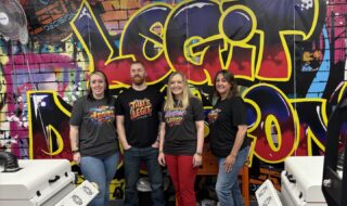 Four people stand together in a print studio with a vibrant graffiti wall behind them, wearing matching 'That's Legit' shirts.