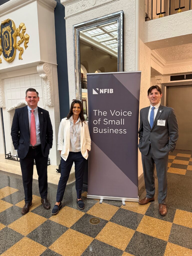 Biryla- - NFIB Three business professionals pose beside an NFIB banner reading 'The Voice of Small Business' in a decorative lobby.