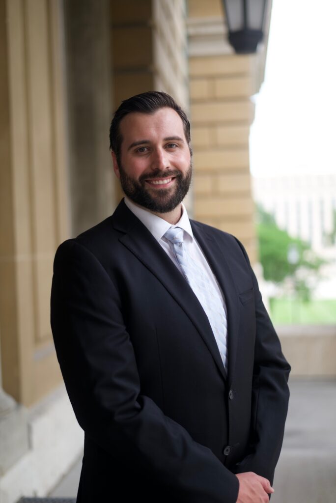 CodyCarlsonIA - NFIB Man in a black suit and light blue tie smiles while posing outdoors beside a stone building.
