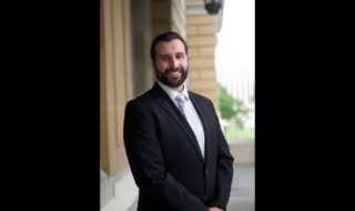 Smiling man in a dark suit and light blue tie standing outside a beige stone building with columns.