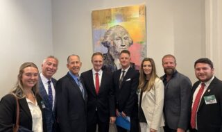 Seven professionals in business attire posing for a photo in a hallway with a colorful abstract portrait behind them.