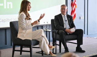 Panel discussion: woman in a light suit speaks with hand gesture to a man in a dark suit; water bottles on the table, American flag in background.