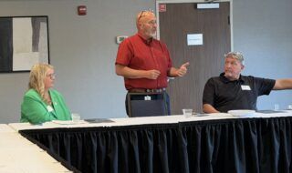 Man in red shirt standing and speaking at a long, white-table conference setup with two seated colleagues nearby; door labeled Mechanical in the background.