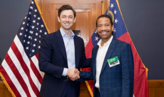 Two men in suits shake hands in front of American flags at a formal event, both smiling.
