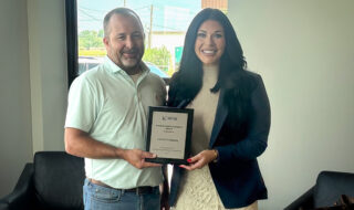 Two adults pose with an award plaque in a bright office lobby, smiling at the camera.