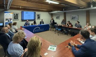 Business meeting in a conference room with a speaker at the front beside a projector screen and Blackstone Valley Chamber of Commerce banner.