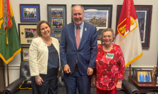 Three smiling adults stand together in an office, flanked by flags and framed photos on the wall behind them.