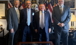 Four men in business suits posing for a photo in a government office, with U.S. flags and a framed seal visible behind them.
