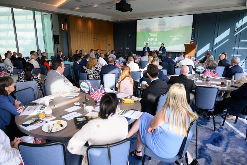 NFIB June Fly-In 2026 Day 2 _ 15 - NFIB Busy conference room with attendees seated at round tables, watching two speakers on stage and a large screen showing 'NFIB DC Fly-In 2026' and Capitol image.