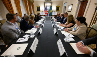 Long rectangular conference table with attendees on both sides, flags at the end, and name placards for a formal meeting in a stately room.