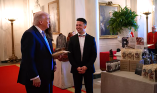 An older man in a dark suit and blue tie shakes hands with a younger man in a black tuxedo on a red carpet in a formal hall with artwork and lamps in the background.