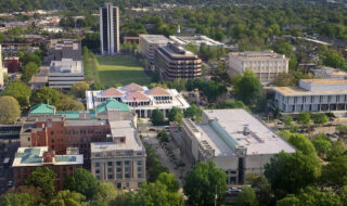 Aerial view of a university campus with a green central quad, surrounding modern buildings and tree-lined streets in daylight.