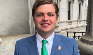 Smiling man in a blue suit and green tie stands outdoors in front of a stone building.