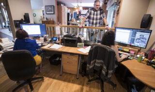 Open office scene with two workers at desks and large computer monitors, a man stands by a kitchen railing in the background.