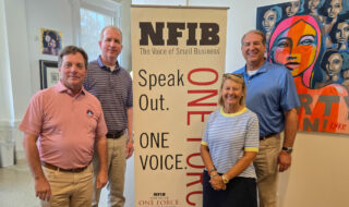 Four adults pose for a photo beside an NFIB banner that reads 'The Voice of Small Business' with 'One Voice' vertically, in a room with a colorful mural on the right.