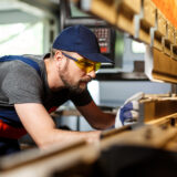 Worker in a blue cap and yellow safety glasses leaning over a workbench with wood planks in a workshop.