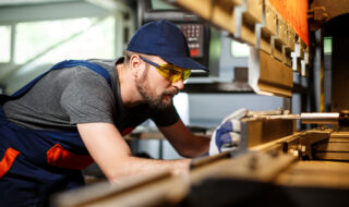 Worker in a blue cap and yellow safety glasses leaning over a workbench with wood planks in a workshop.