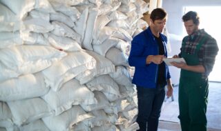 Two men in work clothes review a clipboard next to tall stacks of white bags in a warehouse.