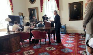 Man at a wooden lectern speaks to a press corps in a formal room with red drapes and portraits on the walls.