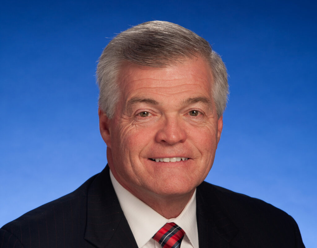 109th General Assembly - Senator Jim Tracy - NFIB Close-up of a smiling man in a dark suit and red plaid tie, against a bright blue sky background.