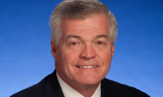 Close-up of a smiling man in a dark suit and red plaid tie, against a bright blue sky background.