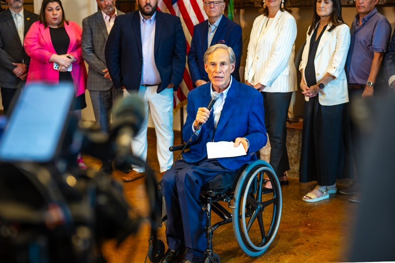 Small Business Roundtable Abbott 1 - NFIB Man in a wheelchair speaks into a mic at a press event, flanked by a diverse group of people and an American flag behind him.