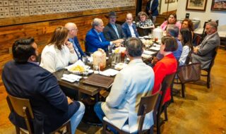 Businesspeople seated around a long table in a restaurant, discussing over lunch with plates and drinks nearby.