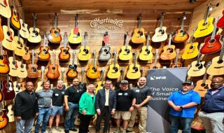 Group of people posing in a guitar showroom with rows of acoustic guitars on wooden walls behind them, including a visible Martin & Co sign.